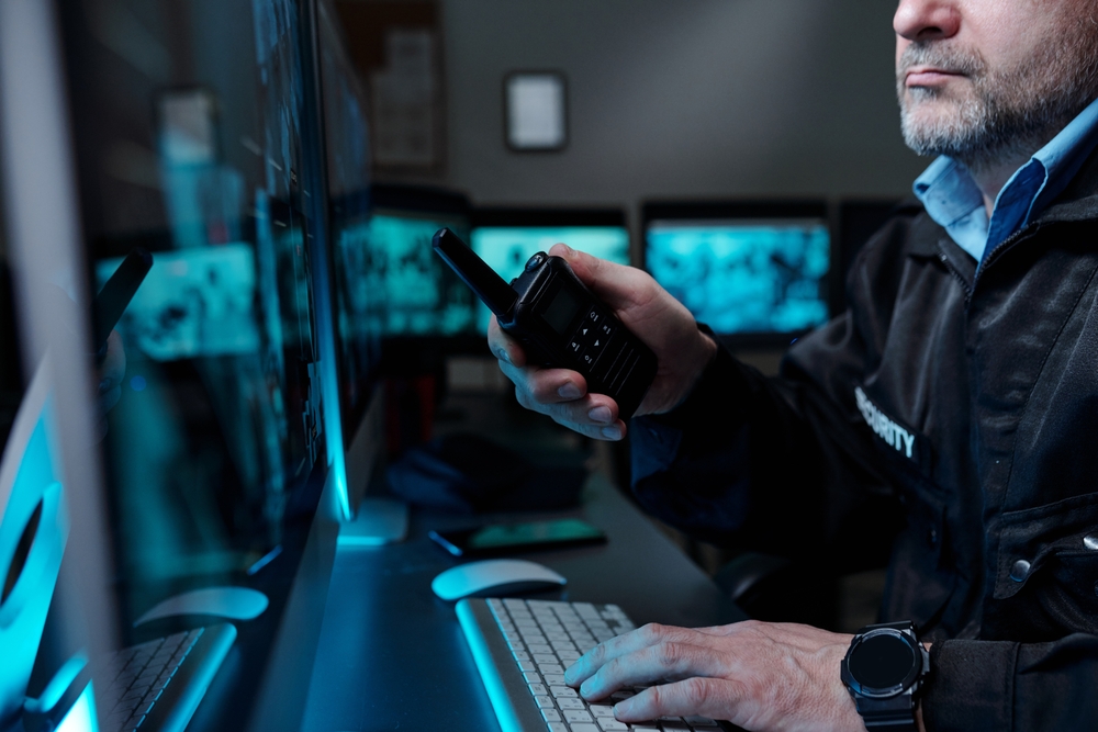 A male security guard for Royal Caribbean Cruises sits in a control room, looking at video footage of an onboard disruption, and calls for backup on his walkie-talkie.